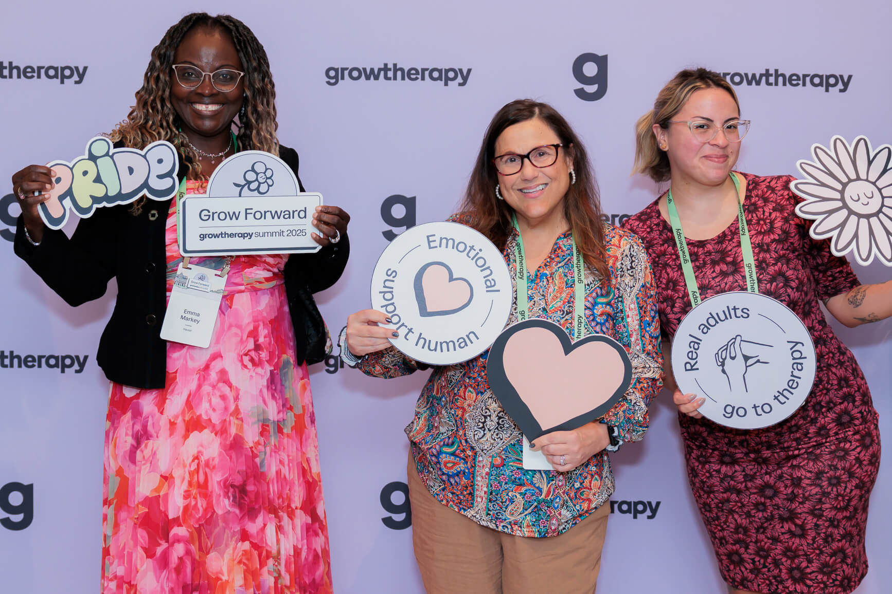 A group of Grow providers pose for a photo, holding up various Grow-branded props, including Pride, the Grow Forward logo, Emotional Support Human with a heart, a separate heart, Real Adults Go to Therapy with an illustration of clasped hands, and a flower with a calm face.