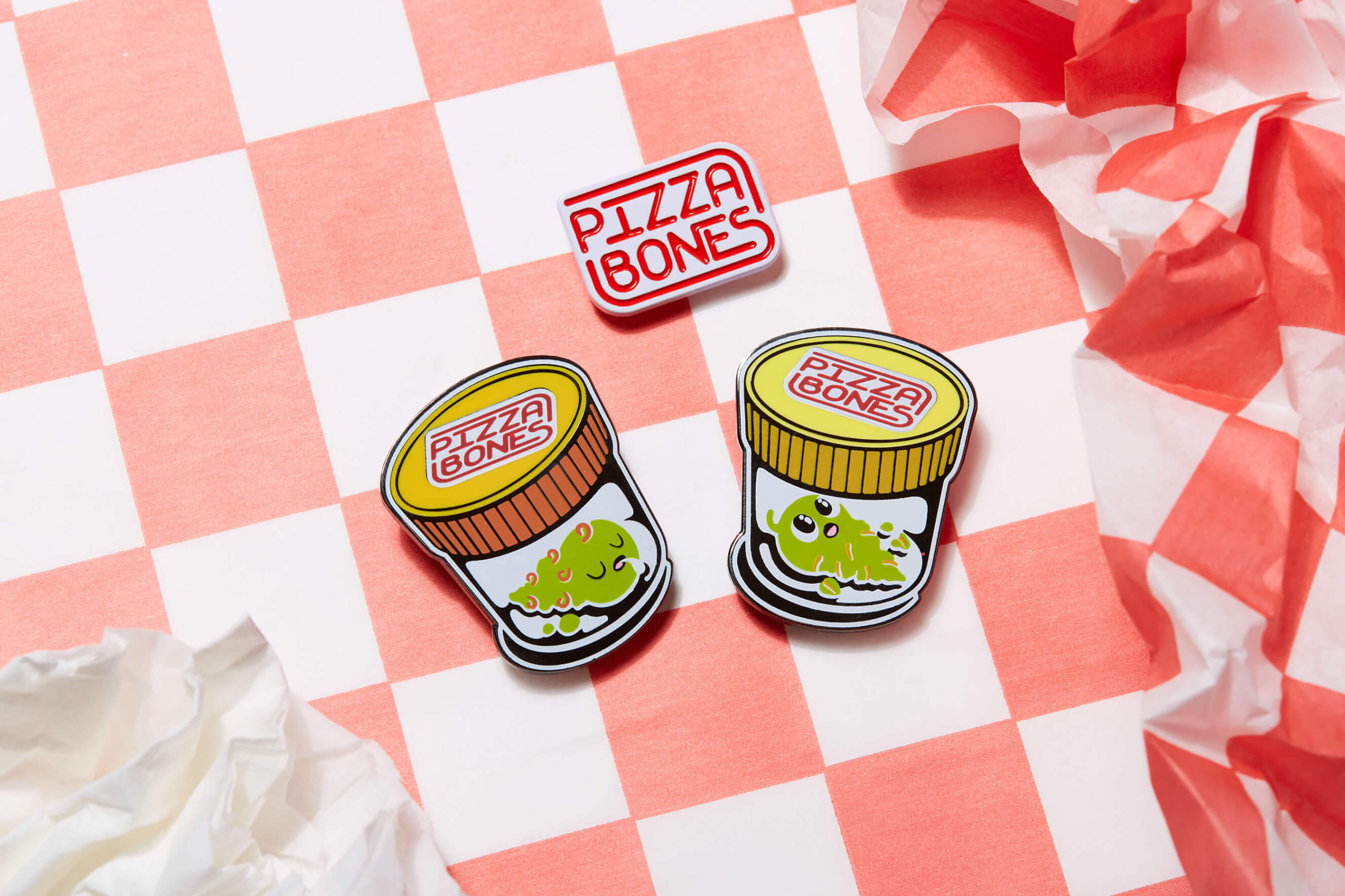 Photo of three Pizza Bones pins, Indica, Pizza Bones Logo, and Sativa. The background is checkered red and white, and there's a crumpled napkin nearby.