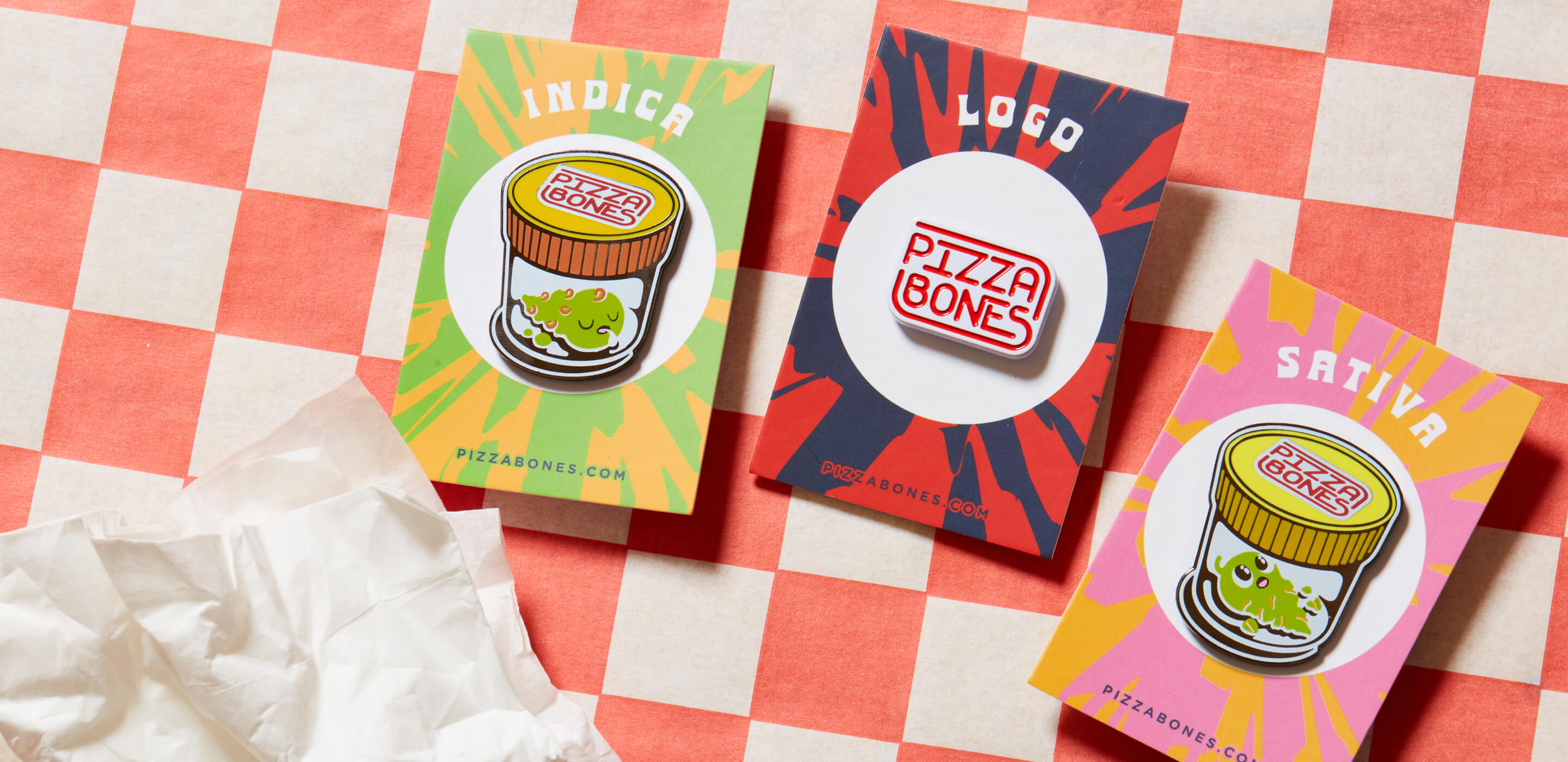Photo of three Pizza Bones pins, Indica, Pizza Bones Logo, and Sativa, sitting on a backing card. The background is checkered red and white, and there's a crumpled napkin nearby.