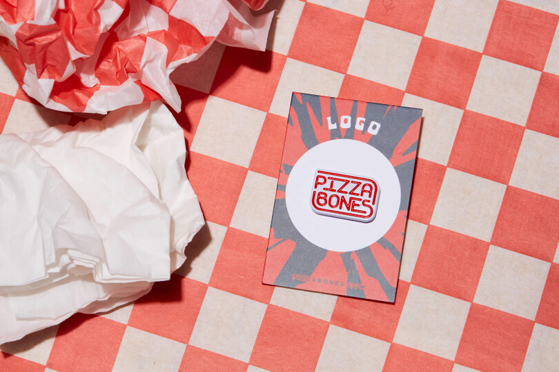 Photo of Pizza Bones Logo pin. The background is checkered red and white, and there's a crumpled napkin nearby.
